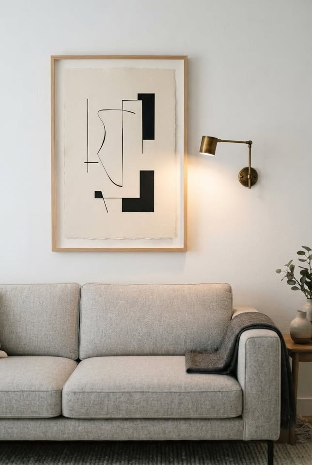 Minimalist abstract art print in a light wood frame hanging above a neutral textured sofa with a brass wall sconce.