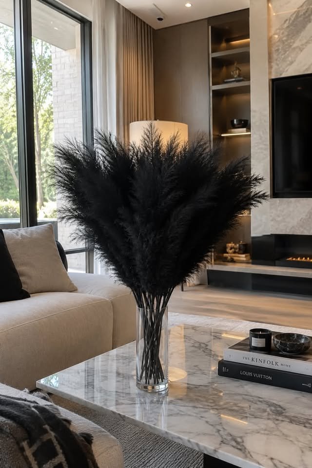 Oversized bundle of fluffy black artificial pampas grass in a clear cylindrical glass vase on a white marble coffee table.