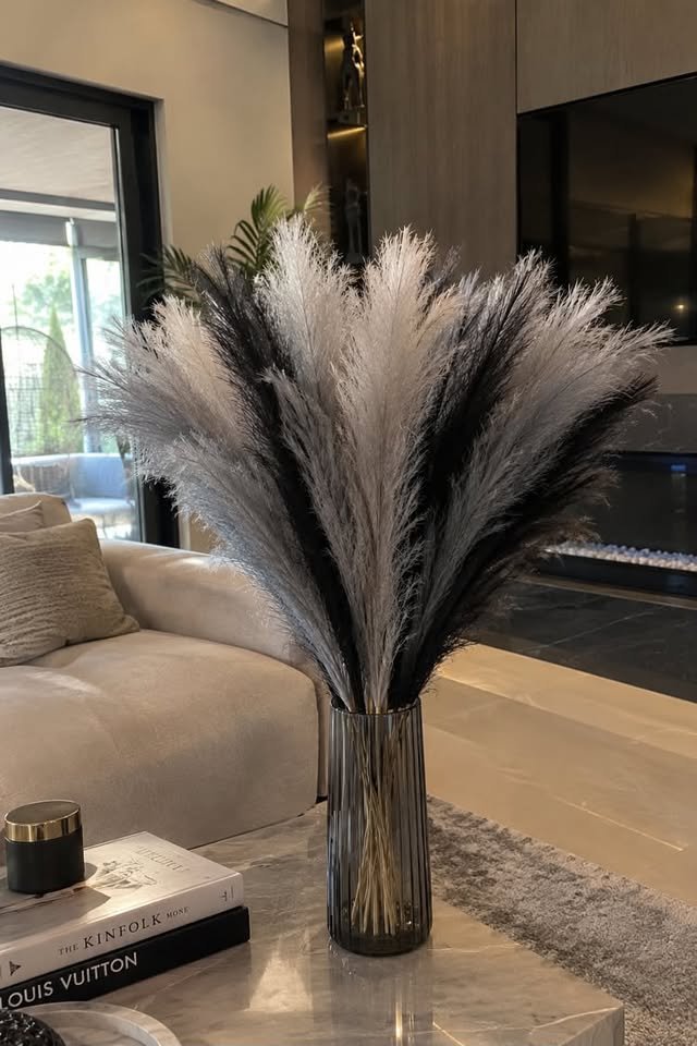 Fluffy black and grey artificial pampas grass stems arranged in a sleek ribbed glass vase on a marble coffee table.