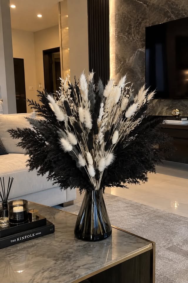 Dramatic mixed bouquet of black faux pampas grass, white bunny tails, and dried botanical stems in a dark glass vase on a marble table.