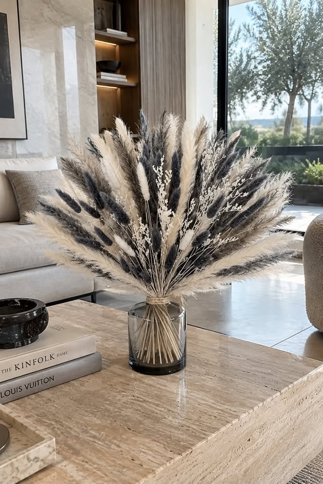 Wide fan arrangement of charcoal grey and cream artificial pampas grass with bunny tails in a short clear vase on a travertine coffee table.