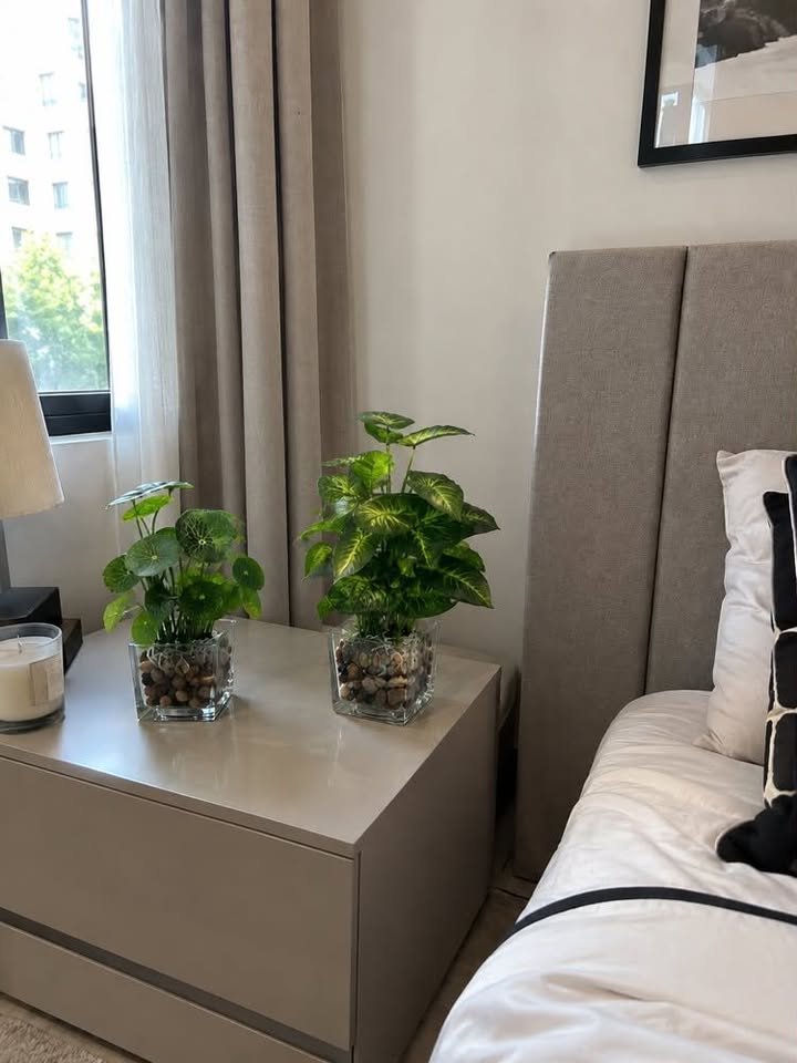 Two artificial green foliage plants in clear glass square vases filled with river pebbles on a neutral bedroom nightstand