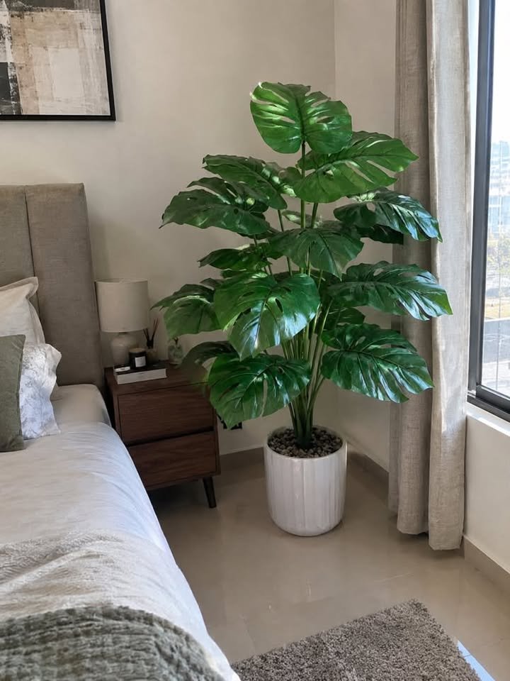 Large artificial Monstera plant in a white fluted ceramic planter filled with stones standing in a bedroom corner beside a nightstand