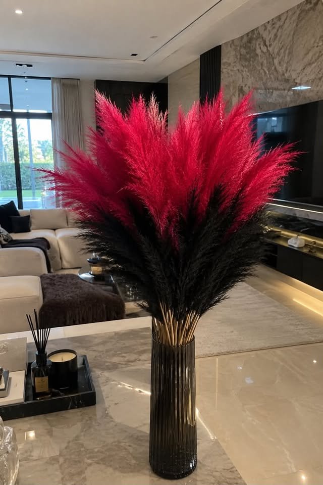  Vibrant hot pink and deep black artificial pampas grass stems arranged in a dark fluted glass vase on a marble tabletop.