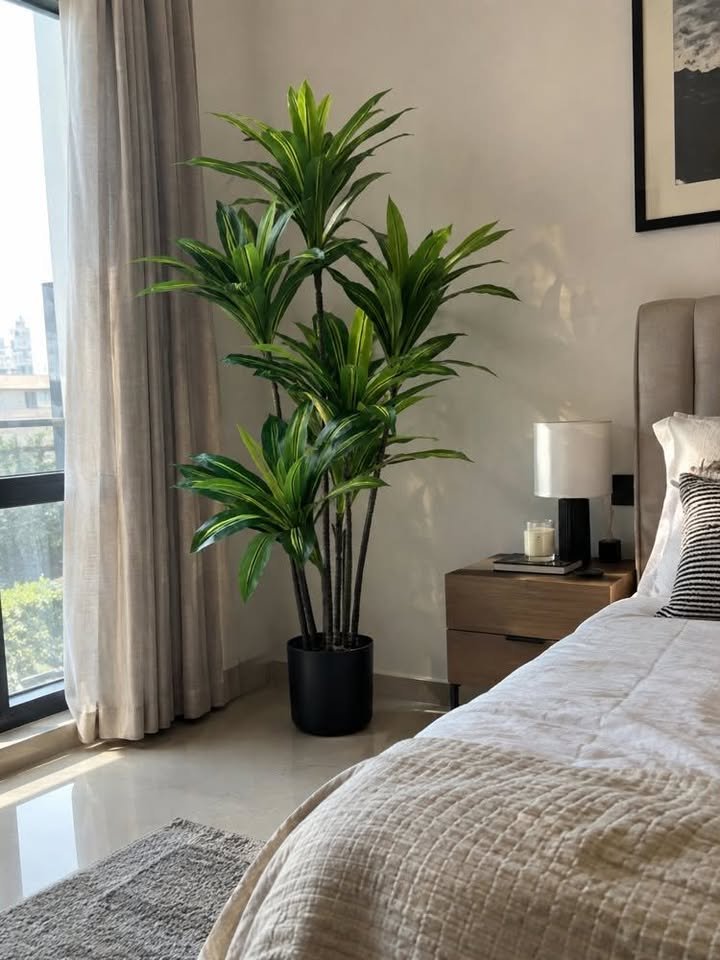 Tall artificial Dracaena Massangeana corn plant in a matte black pot standing in a bright bedroom corner beside a wooden nightstand