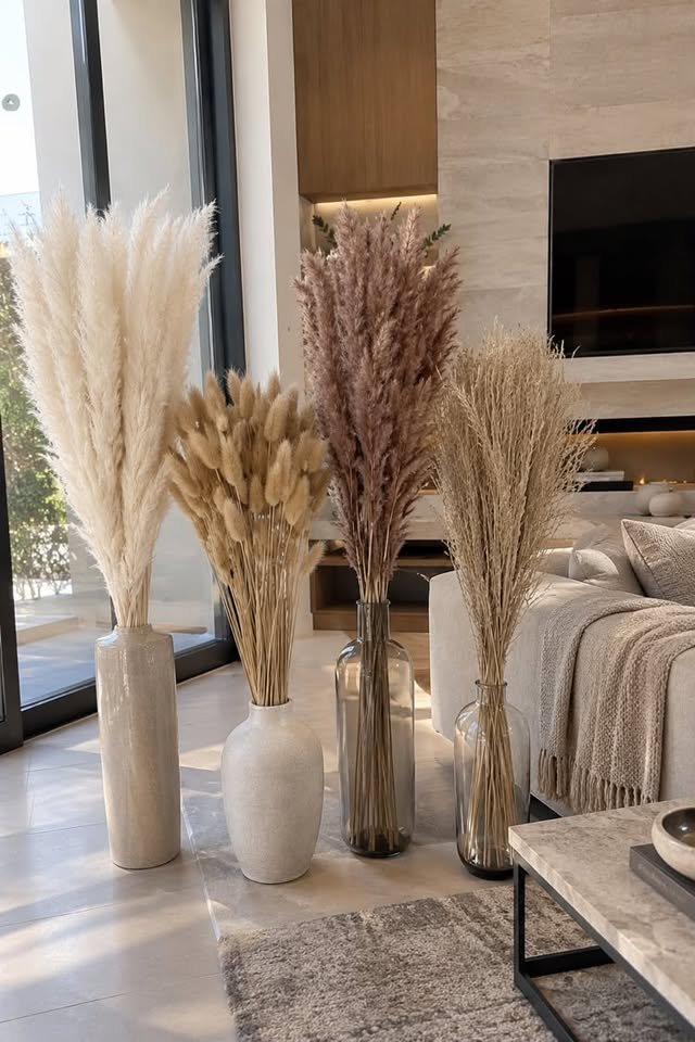 Four floor vases featuring cream faux pampas grass, tan bunny tails, mauve stems, and dried reeds lined up in a bright modern living room.