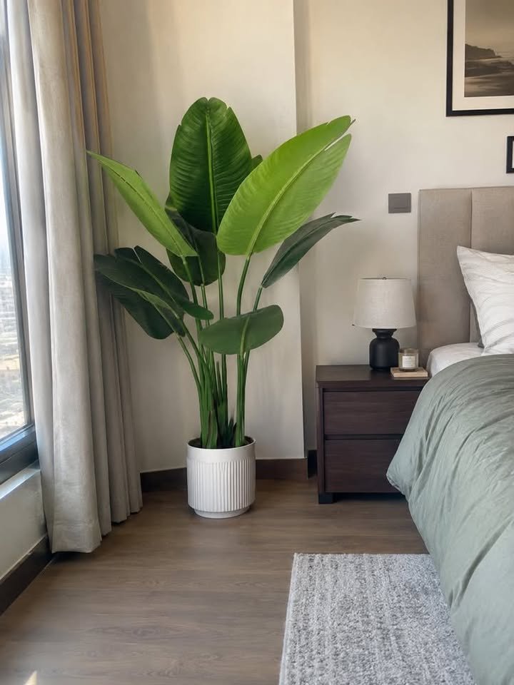 Large artificial bird of paradise plant in a white fluted ceramic pot standing in a modern bedroom corner by the window